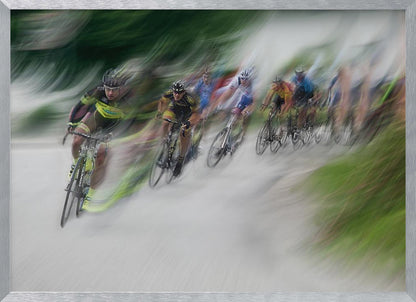 A dynamic, motion-blurred photograph of a group of cyclists in a race, captured with a slow shutter speed to create streaks of color and a sense of high speed. The lead cyclist in a yellow and black jersey is the most defined figure, leaning into a turn on a grey road flanked by blurry green foliage. The entire image is enclosed in a silver frame. Decor