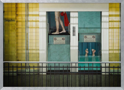 A surreal, framed artwork depicting two teal-colored vertical cabins against a yellow tiled wall. In the left cabin, a person's bare legs are visible as they hold a red towel. In the right cabin, which is filled with water, only a pair of feet can be seen submerged. A black railing is in the foreground, adding to the strange, layered composition. Artwork