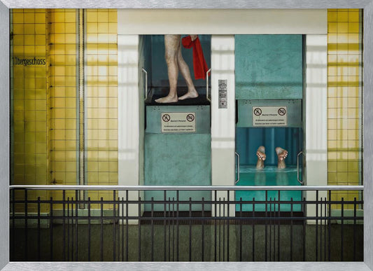 A surreal, framed artwork depicting two teal-colored vertical cabins against a yellow tiled wall. In the left cabin, a person's bare legs are visible as they hold a red towel. In the right cabin, which is filled with water, only a pair of feet can be seen submerged. A black railing is in the foreground, adding to the strange, layered composition. Artwork