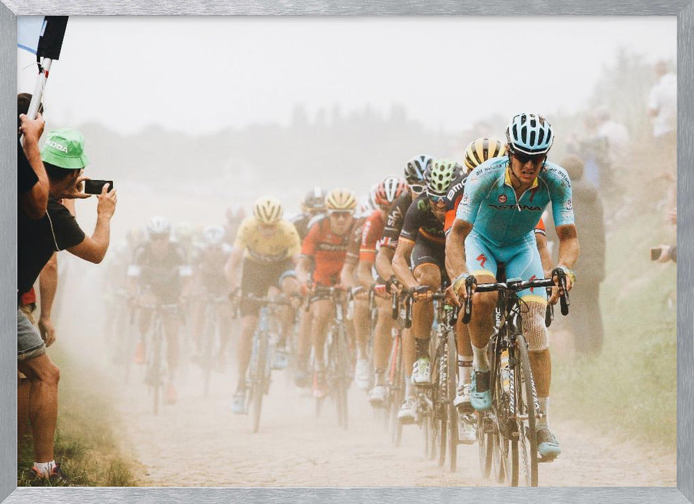 A framed photograph of a peloton of professional cyclists racing down a dusty dirt road. The lead cyclist is in sharp focus, wearing a bright turquoise uniform and a helmet, with other racers in yellow and red jerseys blurred behind him. A spectator on the left is taking a photo with a smartphone. Poster