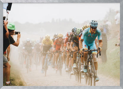 A framed photograph of a peloton of professional cyclists racing down a dusty dirt road. The lead cyclist is in sharp focus, wearing a bright turquoise uniform and a helmet, with other racers in yellow and red jerseys blurred behind him. A spectator on the left is taking a photo with a smartphone. Poster