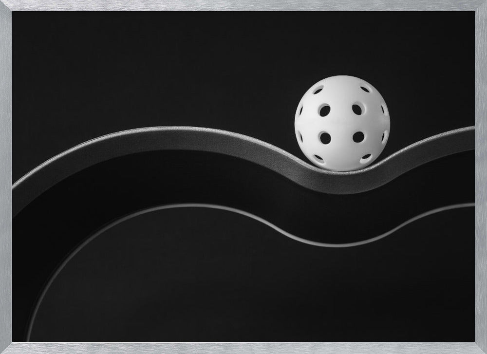 A minimalist black and white photograph of a white pickleball resting perfectly balanced on a thick, wavy, textured line against a solid black background. Below it, a thin white line mimics the curve. The entire piece is framed in silver. Poster