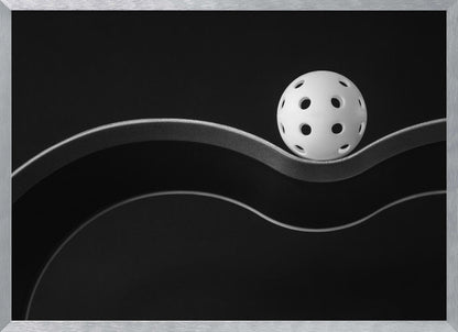 A minimalist black and white photograph of a white pickleball resting perfectly balanced on a thick, wavy, textured line against a solid black background. Below it, a thin white line mimics the curve. The entire piece is framed in silver. Poster