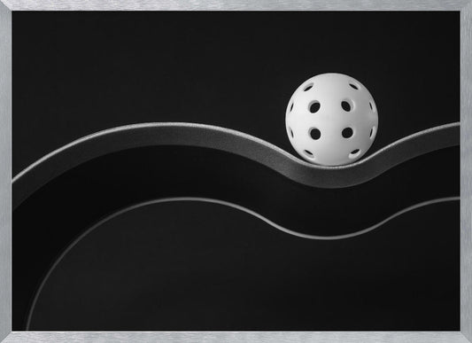 A minimalist black and white photograph of a white pickleball resting perfectly balanced on a thick, wavy, textured line against a solid black background. Below it, a thin white line mimics the curve. The entire piece is framed in silver. Poster