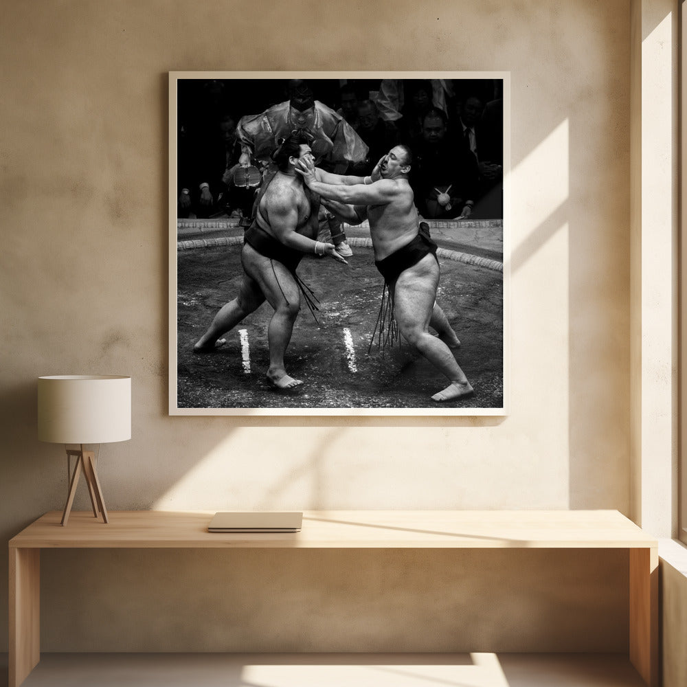 A dynamic black and white action photograph of a sumo wrestling match. Two large wrestlers are in the middle of a clay ring, one pushing forcefully against the other's face. A referee in traditional robes stands behind them, and spectators watch from the background. The entire scene is captured within a light wood frame. Artwork