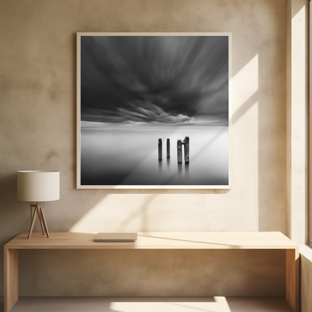 A framed, square, black and white long-exposure photograph of a minimalist seascape. Four dark wooden pilings emerge from still, misty water under a dramatic, streaked sky of dark clouds. Wall Art