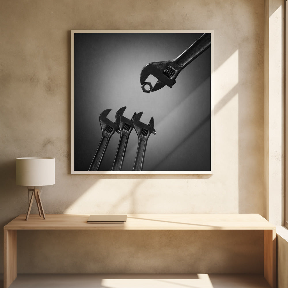 A conceptual black and white photograph in a light wood frame, depicting a large adjustable wrench holding a small nut above three smaller wrenches, which are angled upwards as if they were baby birds waiting to be fed. Print