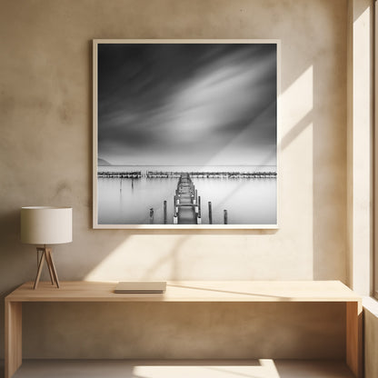 A framed, square black and white long-exposure photograph of a dilapidated wooden pier extending into calm, misty water. The sky is filled with soft, streaked clouds, creating a serene and melancholic mood. Wall Art