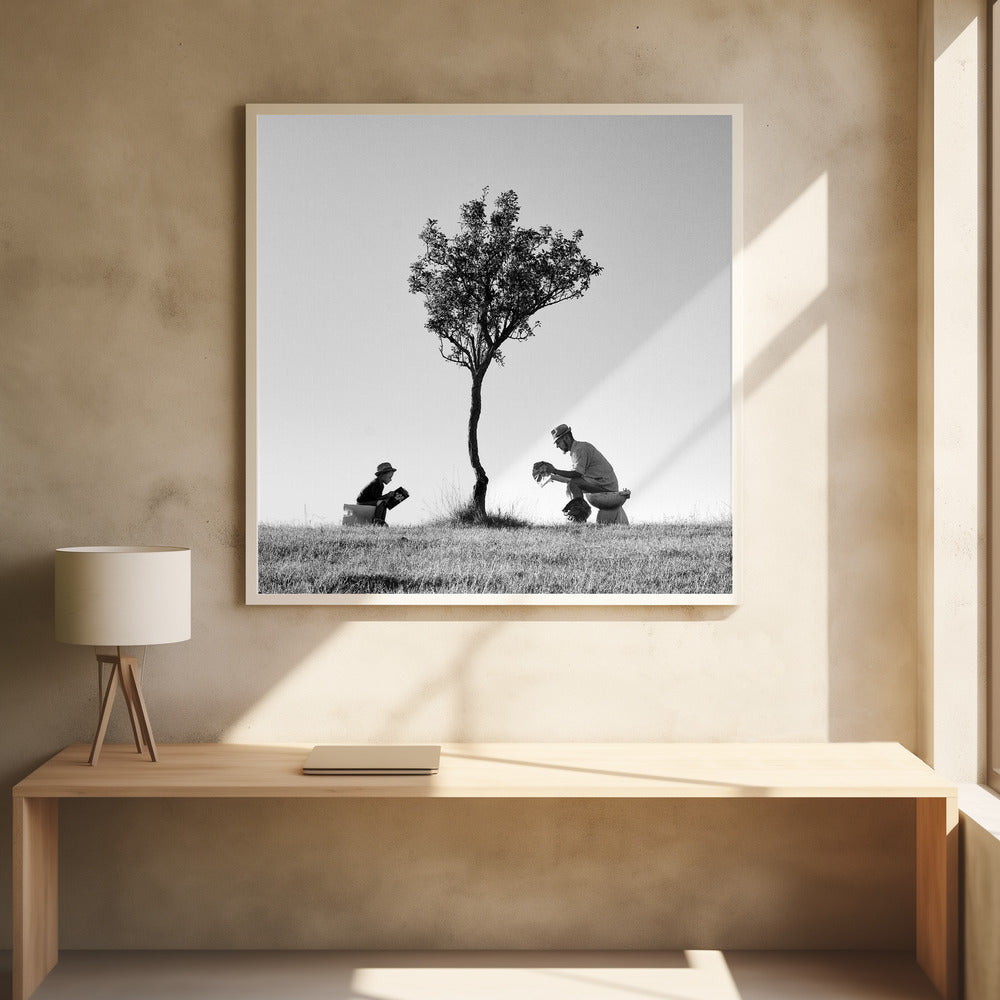 A framed, black and white photograph depicting a surreal scene of a man and a child sitting on toilets in an open grassy field under a single tree, both wearing hats and reading. Print