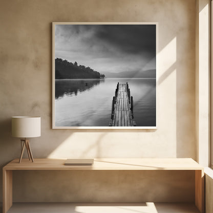 A framed, moody black and white photograph of a wooden pier extending into a calm lake under a cloudy sky, with a forested shoreline and distant mountains in the background. Poster