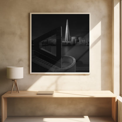 A dramatic black and white architectural photograph of the London skyline at night. The illuminated Shard skyscraper stands tall in the background across a dark river. The foreground features strong geometric shapes, including a large triangular structure and a curved concrete staircase, creating a layered and modern composition. Print