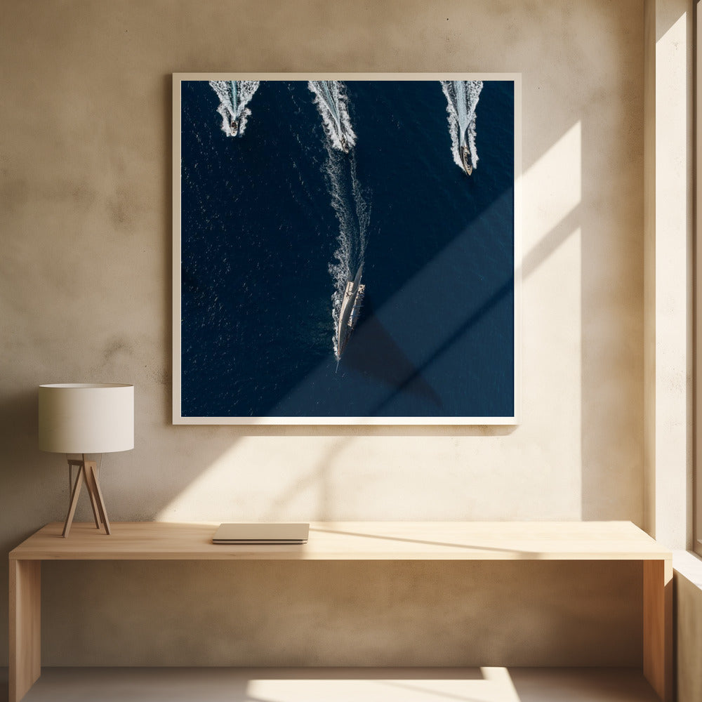 An aerial photograph from directly above shows a large sailboat and three smaller motorboats moving across a deep blue sea, leaving white wakes behind them. The sailboat is in the foreground, casting a long shadow, while the smaller boats are positioned further back. The entire image is enclosed in a light-colored wooden frame. Wall Art