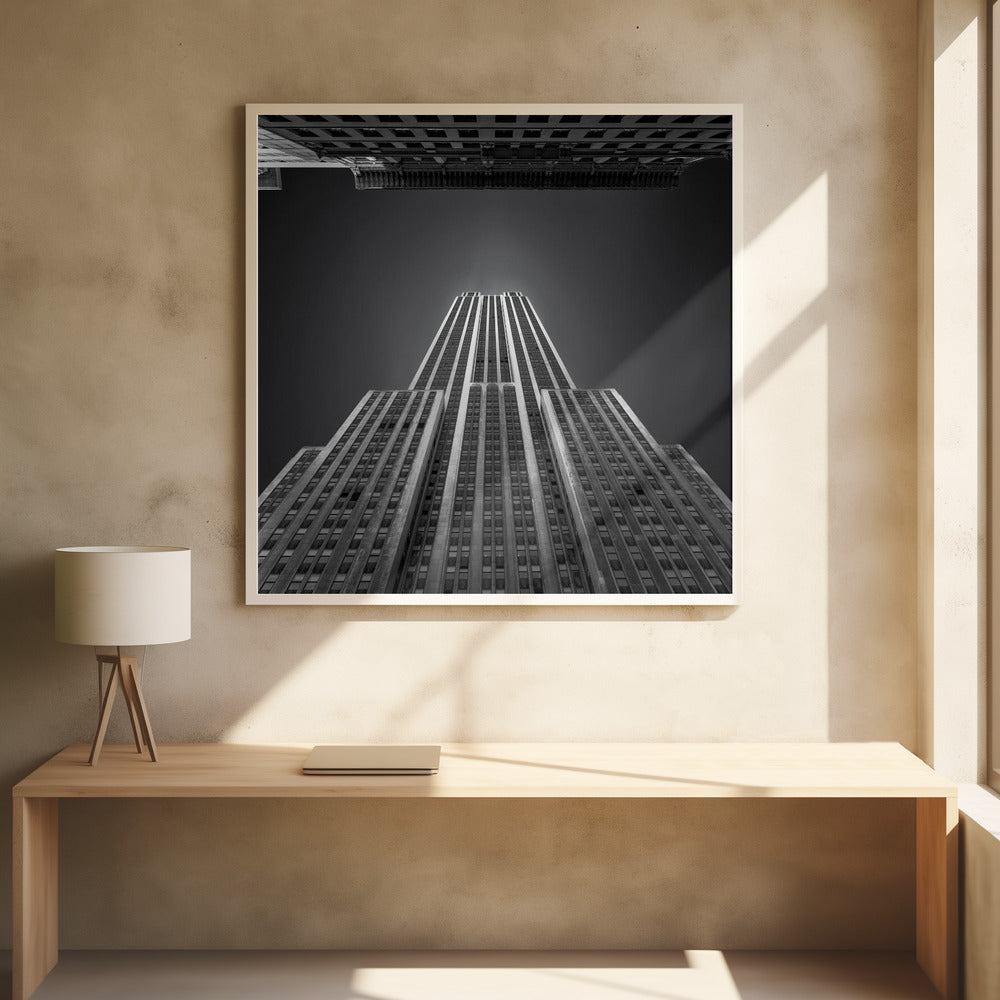A dramatic, low-angle, black and white photograph of the Empire State Building, looking up from the street. The building's Art Deco lines stretch towards a dark grey sky, with another building visible at the very top of the frame, all enclosed in a light brown wood frame. Wall Art
