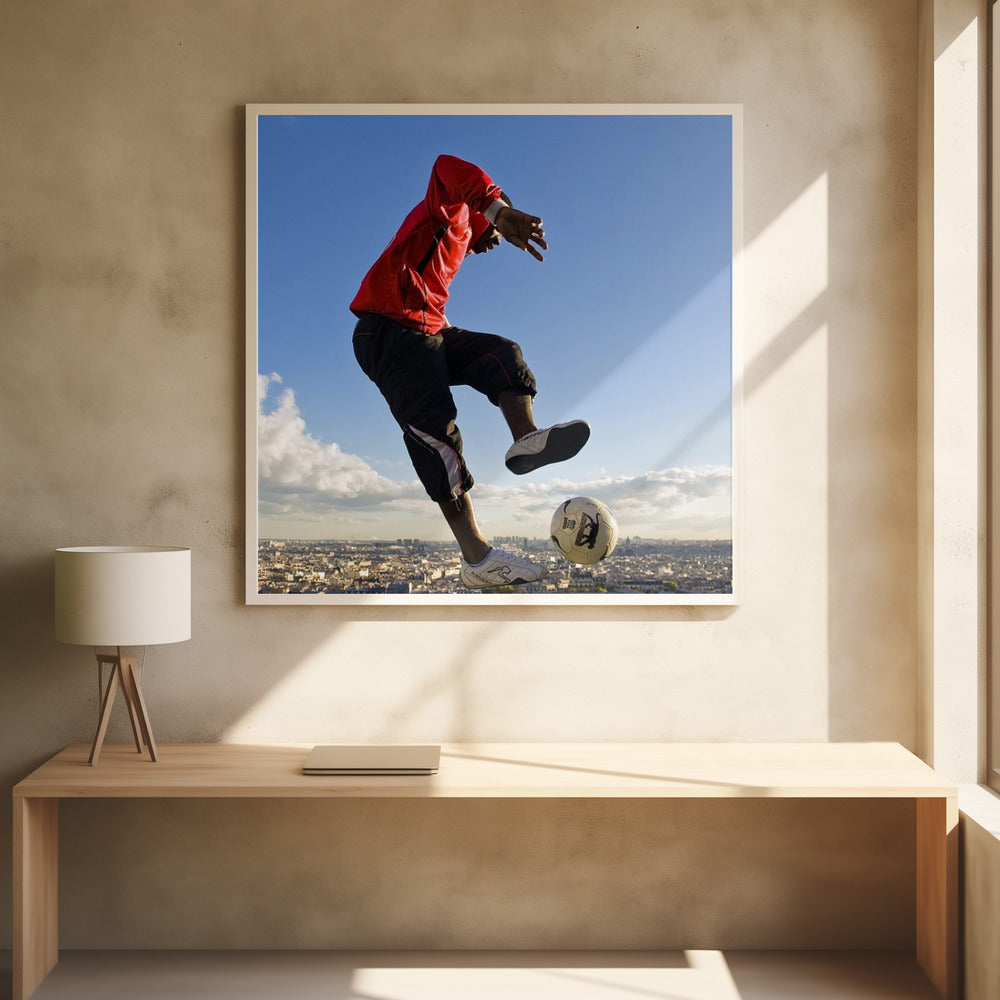 A dynamic low-angle shot of a person in a red jacket and black pants skillfully juggling a soccer ball, suspended in mid-air high above a sprawling city skyline under a clear blue sky, all enclosed within a light wood frame. Artwork