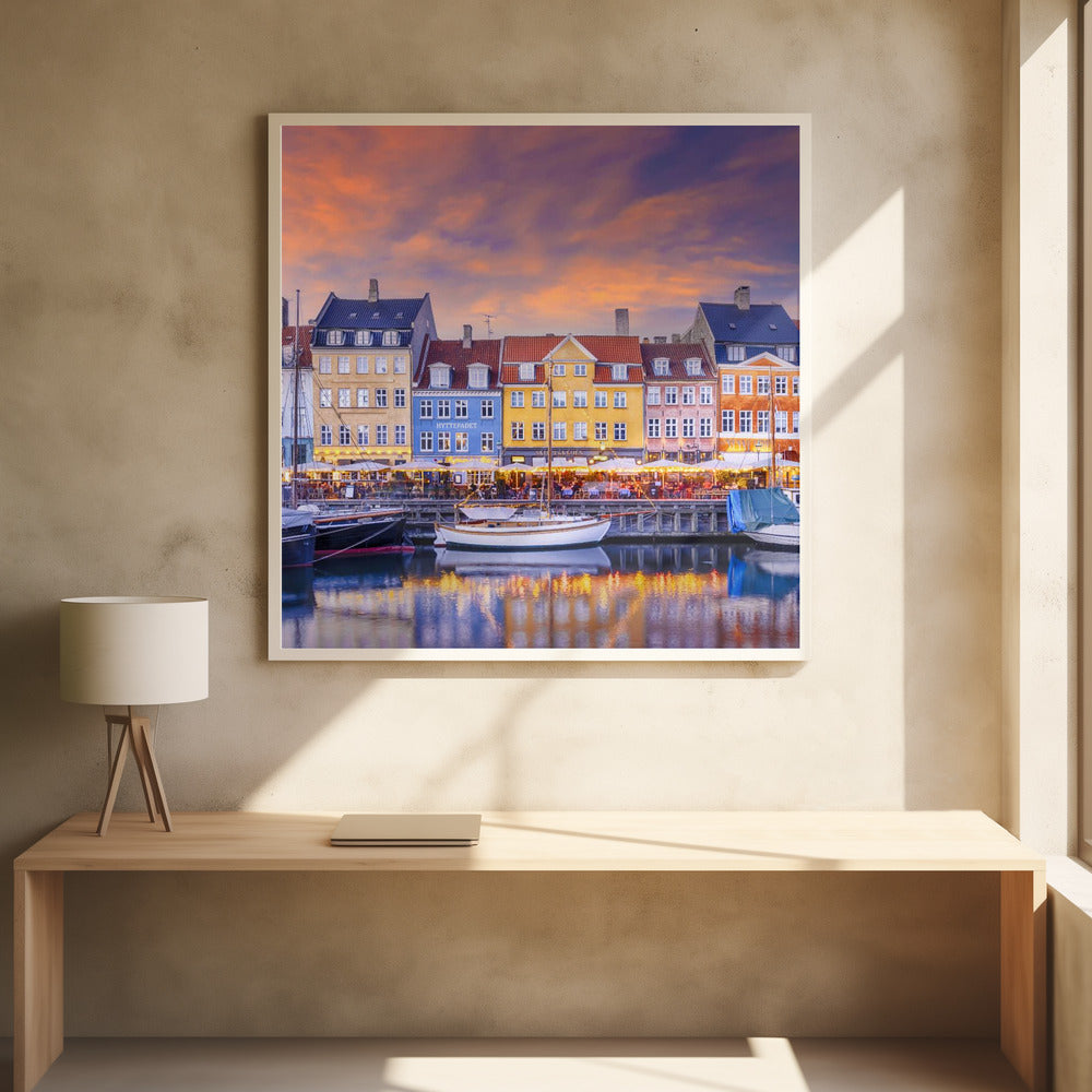 A framed photograph of the Nyhavn harbor in Copenhagen at dusk, featuring a row of brightly colored historic buildings along the canal. Sailboats are moored in the water, which reflects the warm glow of the building lights and the vibrant orange and purple sunset sky. Print