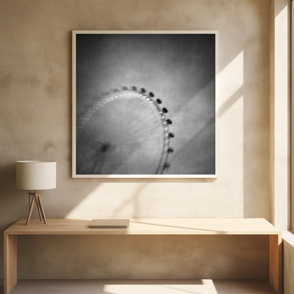 A moody, black and white long-exposure photograph of a large Ferris wheel, creating a soft, blurry effect of its rotation against a grainy gray sky. The artwork is presented in a simple, light wood frame. Poster