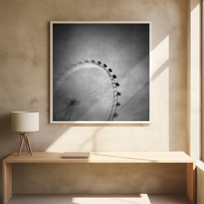 A moody, black and white long-exposure photograph of a large Ferris wheel, creating a soft, blurry effect of its rotation against a grainy gray sky. The artwork is presented in a simple, light wood frame. Poster
