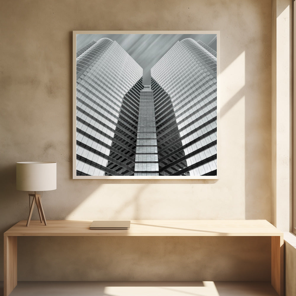 A low-angle, symmetrical black and white photograph of a modern glass skyscraper. The two towers of the building converge towards the center, creating a V-shape against a sky with motion-blurred clouds. The photo is enclosed in a light-colored wooden frame. Print