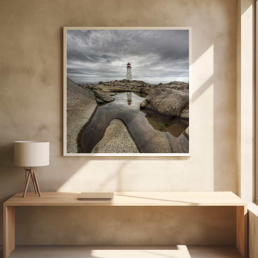 A framed photograph of a white lighthouse with a red top standing on a rocky shore under a dramatic, cloudy sky. In the foreground, a tide pool perfectly reflects the lighthouse. Artwork