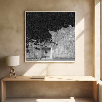A framed, square black and white photograph showing a stark contrast on an ivy-covered wall. The top half is dense with dark leaves, while the bottom half is a lighter, textured wall. A young girl with long hair, seen from behind, stands in a recessed white doorway in the center. Wall Art