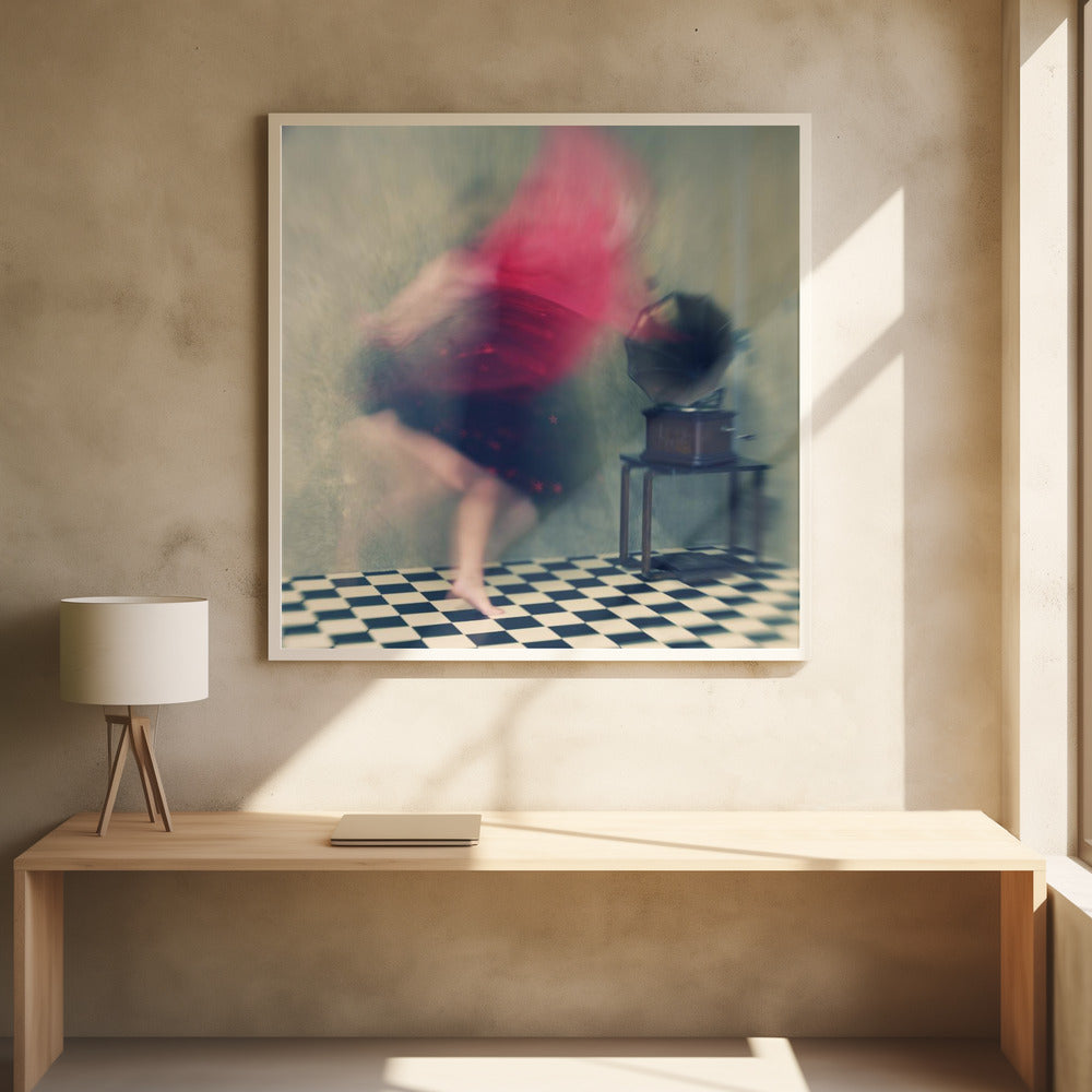 A motion-blurred, artistic photograph in a light wood frame showing a woman dancing barefoot on a black and white checkered floor. She wears a bright red top and a dark skirt, both blurred by her movement. Next to her stands a vintage gramophone on a small table, against a muted, textured wall, creating a dreamy, nostalgic atmosphere. Print